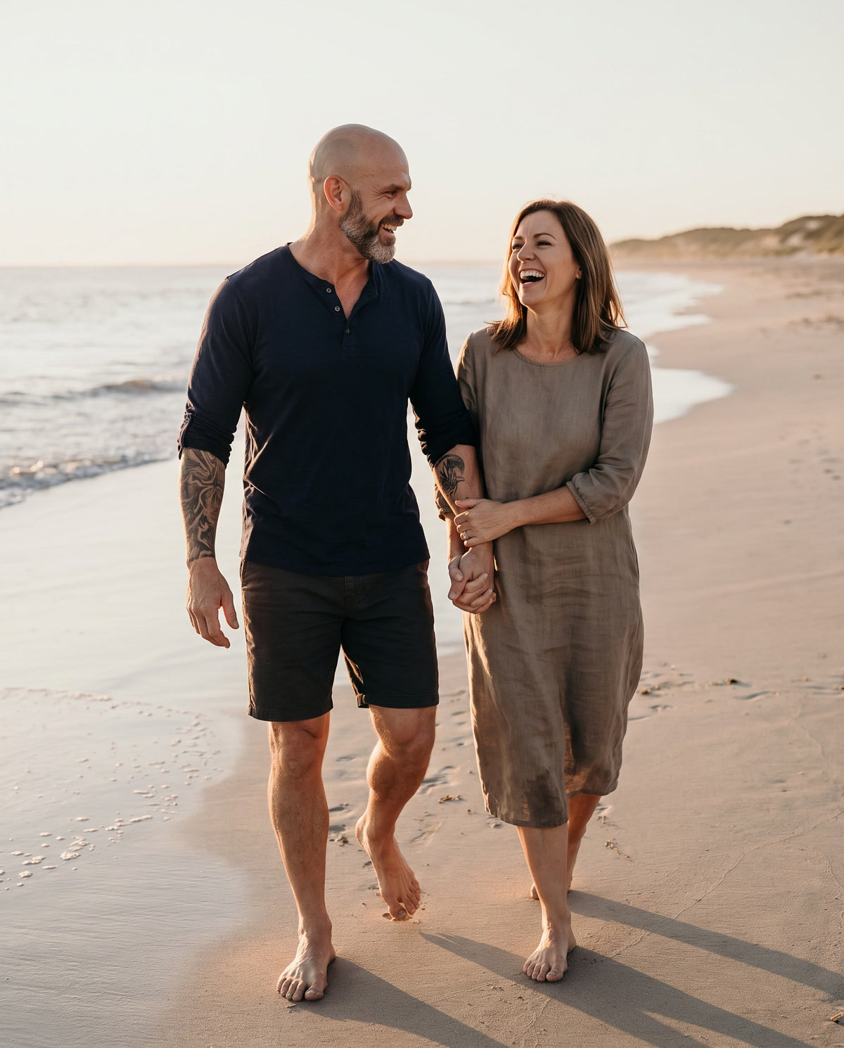 A man and his partner walking barefoot along a beach at sunset, laughing together
