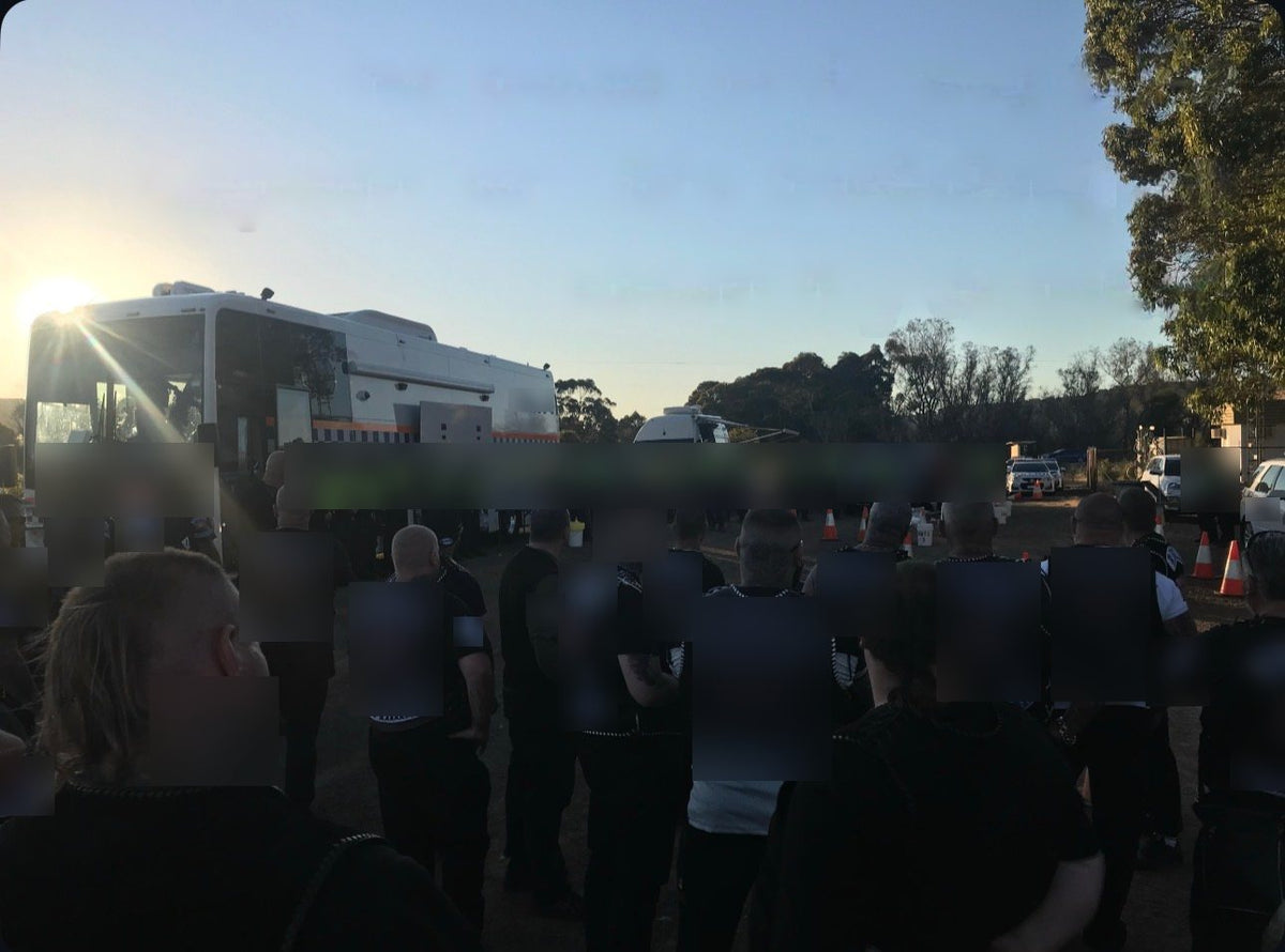 A crowd gathered at sunset beside large vehicles and gum trees; identifying patches, faces and markings are blurred.