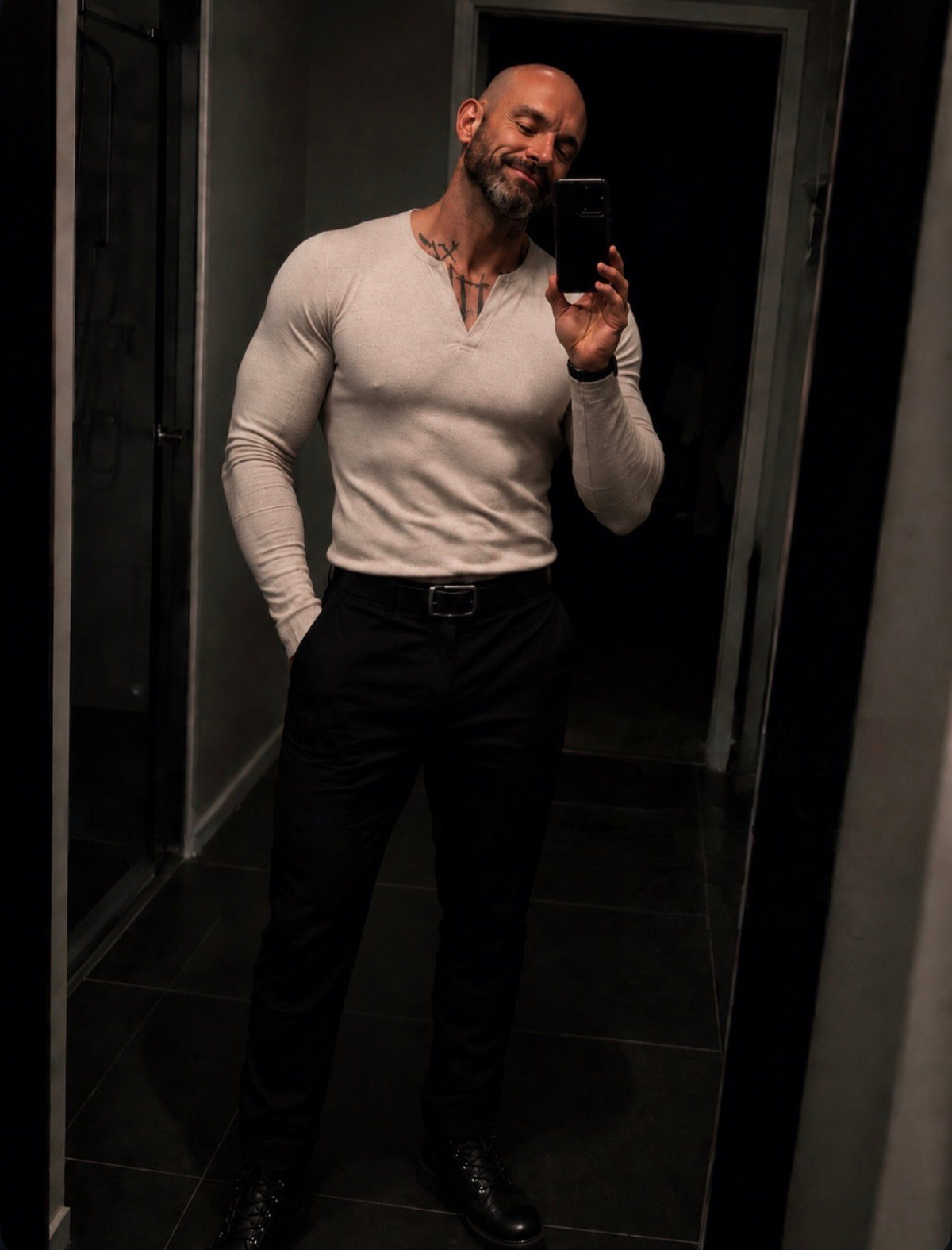 Mirror selfie in a dark hallway, smiling with eyes closed, in a fitted cream henley, black trousers and boots.