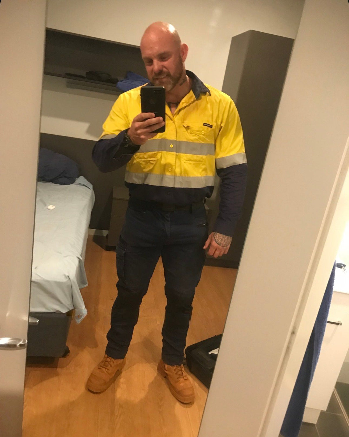 Mirror selfie in a bedroom, wearing a yellow-and-navy hi-vis work shirt, navy work trousers and tan work boots.