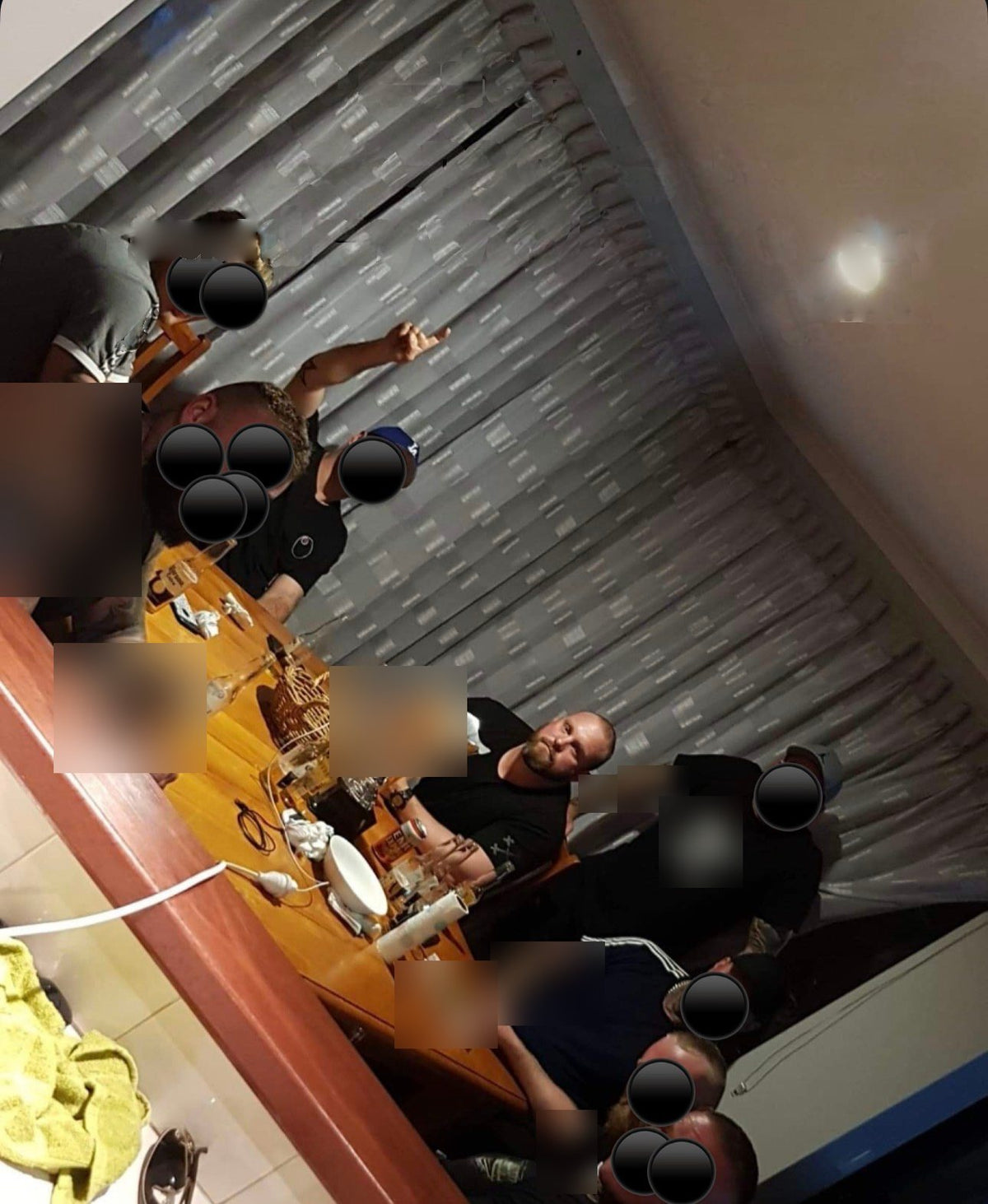 A group gathered around a kitchen table at night; faces, tattoos, table items and logos are blacked out or blurred.
