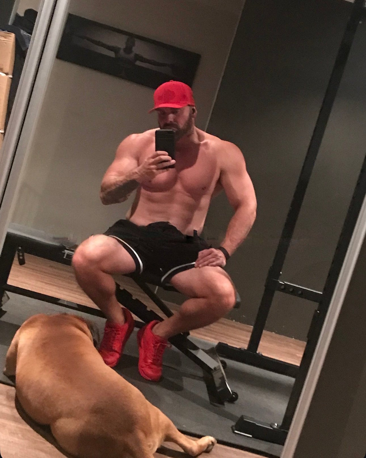 Sitting on a weight bench in a home gym in black shorts and red shoes, a dog lying on the floor.