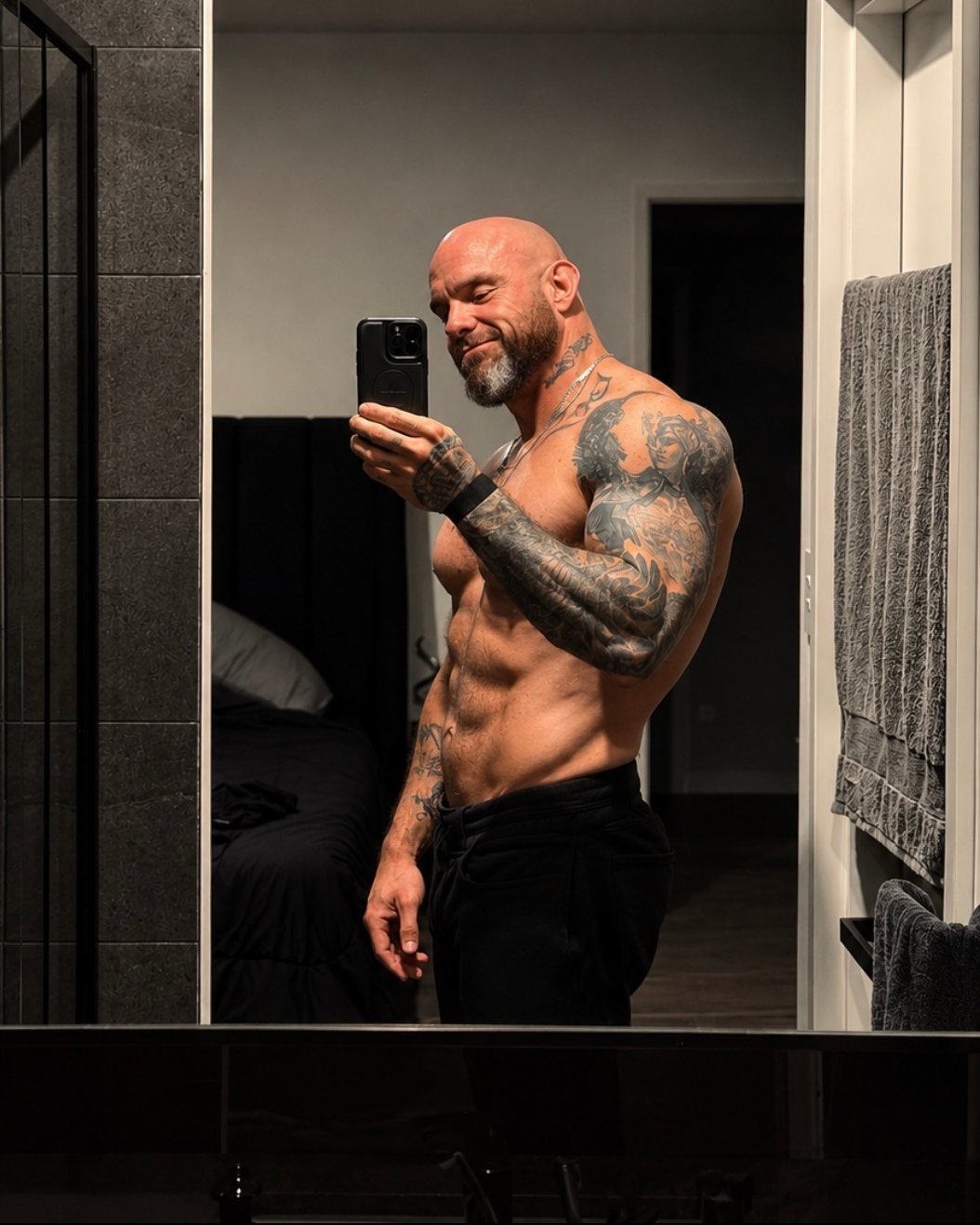 Willow, shirtless and tattooed, taking a mirror selfie in a dark bathroom.