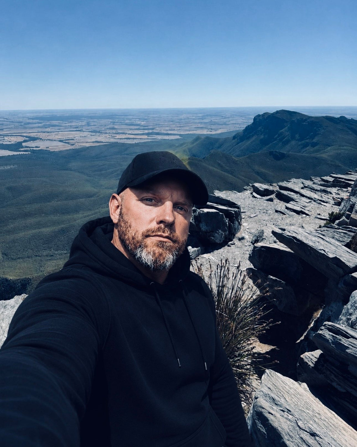 Selfie at a rocky mountain summit in a black cap and hoodie, with a wide valley and farmland stretching to the horizon behind.