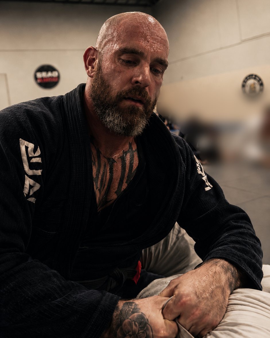 Willow in a black gi, sweating, gripping a training partner's sleeve on the mats; other trainees blurred in the background.