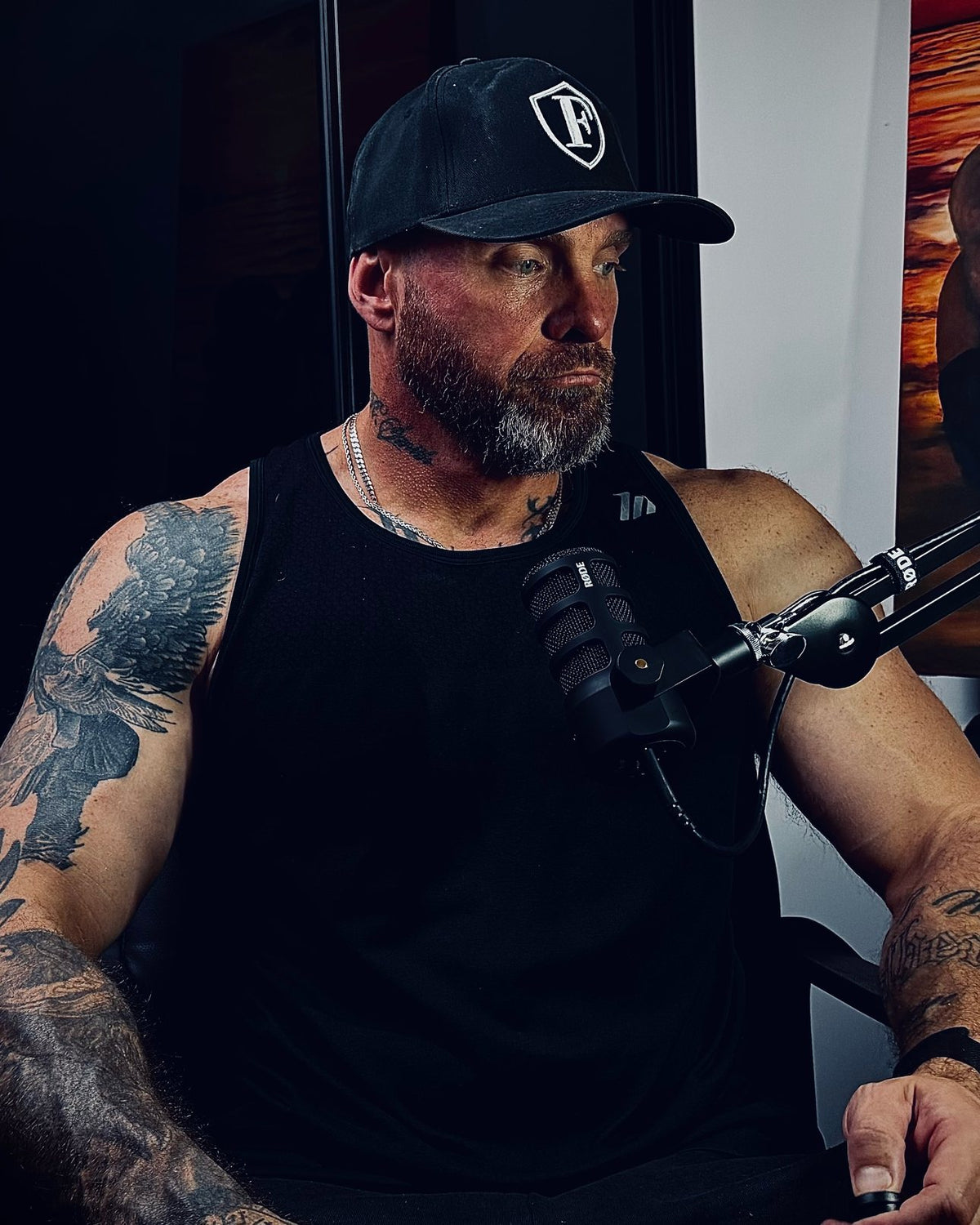 Seated at a podcast microphone in a black cap and black singlet, tattooed arms, a painting on the wall behind.